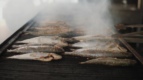 Sea fish is fried on a baking sheet in a hotel or restaurant kitchen. Chef hand with rubber glove turns the fish over with culinary tongs through steam. Close up 