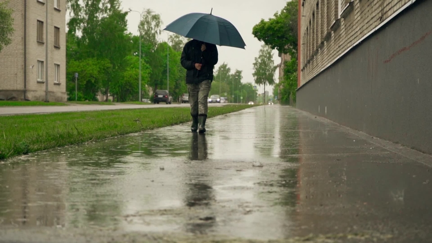 Girl walking in boots on wet ground at rainy day. Steadicam slow motion footage. Tracking female in focus walking on wet surface with reflection of buildings. 