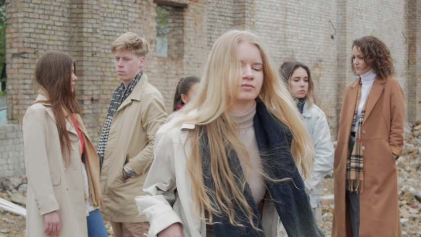 Blonde in a coat performs a dance or expresses nervousness and stops among a group of young people on the ruins. The youth makes a theater etude amid a collapsed brick building Royalty-Free Stock Footage #1031473946