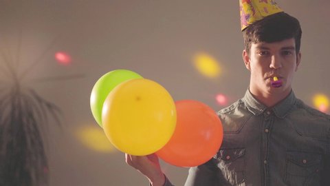 Portrait of young man in birthday hat with noisemaker in mouth looking in the camera holding balloons. Bright lights of different colors on his face and on the wall. The guy at the party, celebration