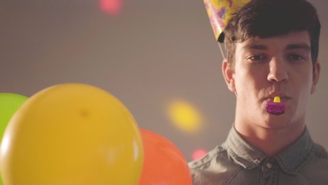 Portrait of young cute man in birthday hat with noisemaker in mouth looking in the camera holding balloons. Bright lights of different colors on his face and on the wall. The guy at the party