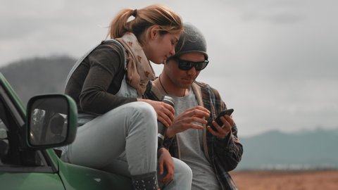 Two travelers at the SUV during a car summer trip. A woman sitting on the hood with a thermocup, a man uses a mobile phone, looking for a route, talking, smiling. Travelers on a halt. Slow motion, 4K