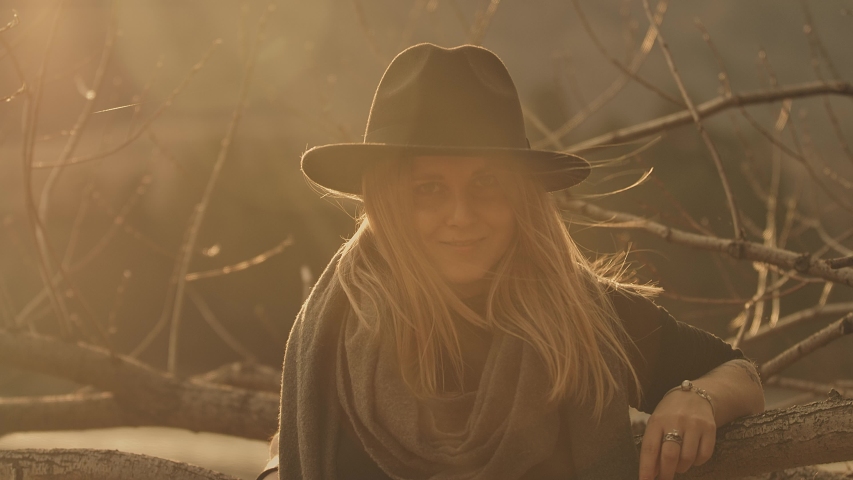 Close-up portrait: a beautiful young woman in the light of the sunset in a black hat standing in the open, looking at the camera and smiling, hair develops wind. Golden sunlight, slow motion, 4K