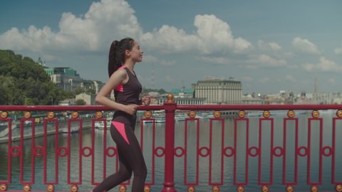 Side view of athlete fit asian woman with armband running across bridge over cityscape background. Self motivated sporty fitness female jogging outdoors while doing morning workout.