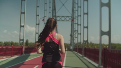 Back view of young brunette sportswoman with armband doing sports training outdoors. Fitness female jogger in tight sport outfit running across pedestrian bridge, active, healthy lifestyle concept.