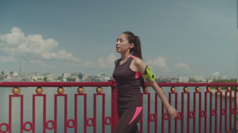 Side view of deep breathing female runner with armband walking along river bridge. Chinese athletic female in sporty outfit calming breathing after running, active, sport, healthy lifestyle concept.