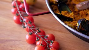 On wooden table tomato cherry moving camera slider showing hand of chef decorating paella with prawn, slow motion close up video, ripe ingredients of prepared served paella spanish traditional cuisine