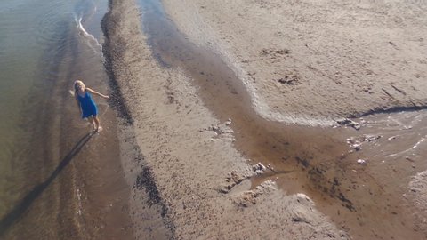 Top view aerial shooting from flying drone of a blonde in a blue dress walks on the water on the beach of the Baltic Sea, early in the morning. Sunny day. Long shadows.