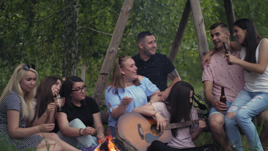 Group of young friends is singing songs around a campfire in the forest, playing the guitar and roasting marshmallow. Green trees and grass are visible. Tourism, teen life, adventure concept | Shutterstock HD Video #1031515352