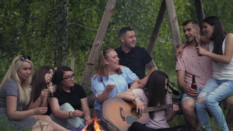 Group of young friends is singing songs around a campfire in the forest, playing the guitar and roasting marshmallow. Green trees and grass are visible. Tourism, teen life, adventure concept