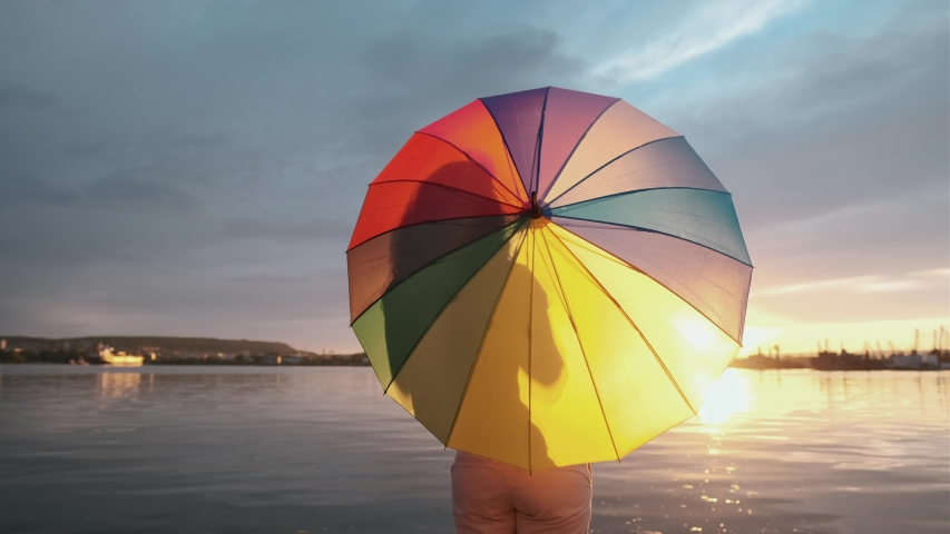 Young woman stands umbrella rotates quay at seaport it against the background of a sunset and a bright sun disk, with a cloudy rainy sky. Rain, clouds, cloudy. Tourism. Freedom