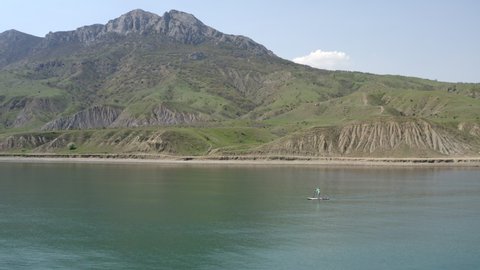 Remote view. Surfer floats on supboard in distance on calm blue sea. View from water to beautiful mountain landscape. Narrow strip beach passes in front mountain. Clear summer day