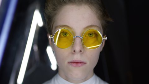 Close up of teenage Caucasian girl with short haircut and no makeup wearing yellow round shaped sunglasses looking at camera against black background with triangular neon lamps
