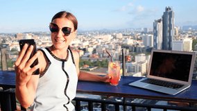 female makes selfie in a cafe on the roof of a skyscraper. girl sitting with a laptop and an orange cocktail on against the panoramic view.