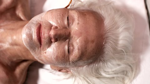 Close-up top face shot of elegant elderly Caucasian woman lying on couch with closed eyes and relaxed smile, and female beautician applying skin moisturizing mask on wrinkly face with cosmetic brush