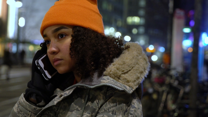 Beautiful biracial African American girl teenager young woman on urban city street at night in winter wearing orange hat and camouflage jacket talking on mobile cell phone