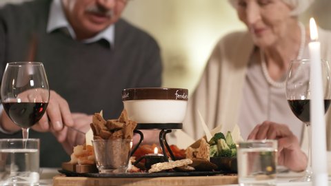 Close-up object shot of cheese fondue starter sitting on table in restaurant, with candles and glasses of red wine beside, and mature man and woman dipping snacks in cheese and eating in background