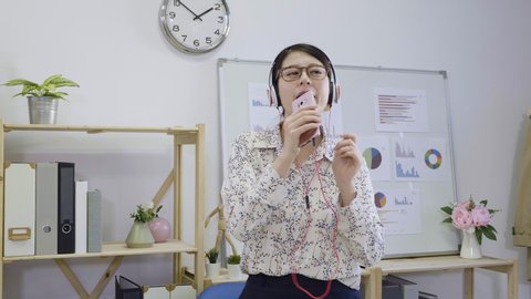 Young joyful woman designer in glasses and headphones listening to music on her smartphone and dancing while sitting at table in office indoors. female worker slide chair to back singing microphone