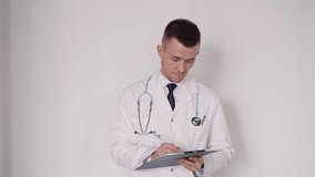 Concentrated, focused and serious smart doctor fills paper form document after visiting new patient. He looking on blank and standing against white wall in bright light interior room