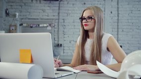 Attractive and charming woman in formalwear sitting behind work table in bright light modern interior. She looking at laptop and speaking in video call inside loft style with natural soft daylight