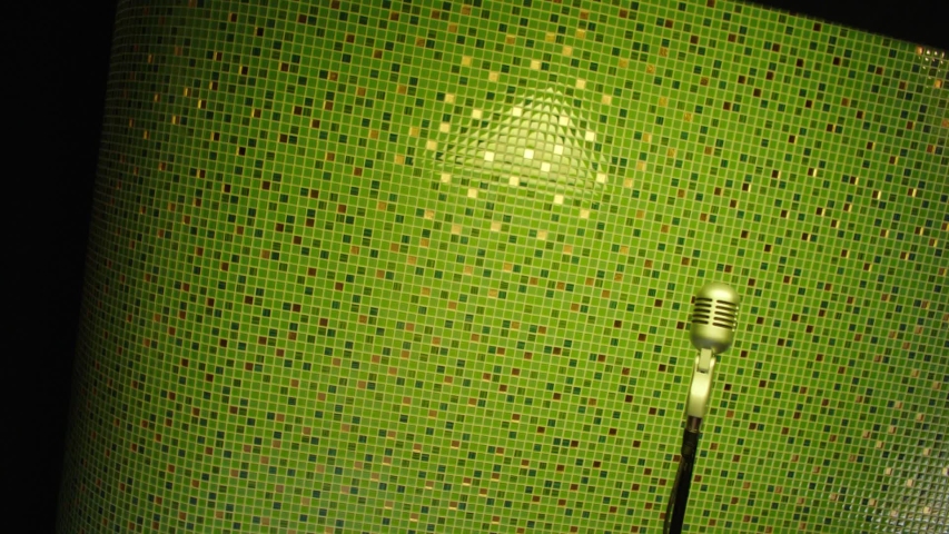 Mature brunette bearded man wearing stylish sunglasses and white t-shirt emotionally dances and sing holding grey stationary microphone in studio with changing color illumination.