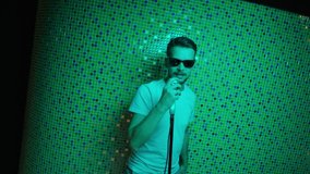 Attractive mature bearded male put on stylish sunglasses and white t-shirt emotionally sings holding grey stationary microphone in studio with rapidly changing colorful light.