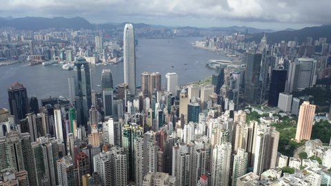 Aerial panorama of Hong Kong viewed from top of Victoria Peak with skyscrapers on HongKong Island & in Kowloon Downtown by Victoria Harbour & residential towers on Mid-Levels hillside under cloudy sky