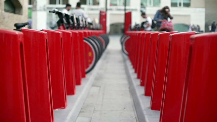 Red bicycle docks in the centre of Barcelona. Wheels and metallic parts of a bike stations. People walking around the square near museum of contemporary art. 