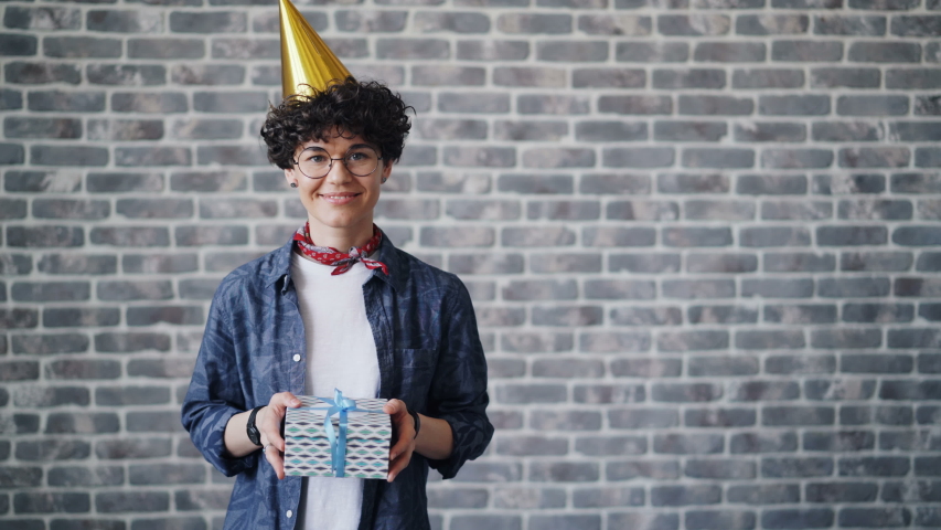 Portrait of cheerful young woman in bright party hat holding gift box and smiling looking at camera standing on brick background. Happy people, presents and holidays concept.
