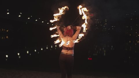 Rear view of young woman artist performing art of juggling fire fans and seductively looking back at viewer during night fireshow on river bank. Pink-haired firegirl spinning burning fans at night.