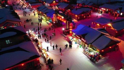 HEILONGJIANG, CHINA – JANUARY 2019: Tilted drone shot of people walking through illuminated snow covered mountain village in remote region of Northern China at night