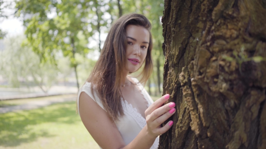 Portrait glamorous young girl with long brunette hair wearing a long white summer fashion dress standing next to a tree in the park. Leisure of a pretty woman looking at the camera outdoors.