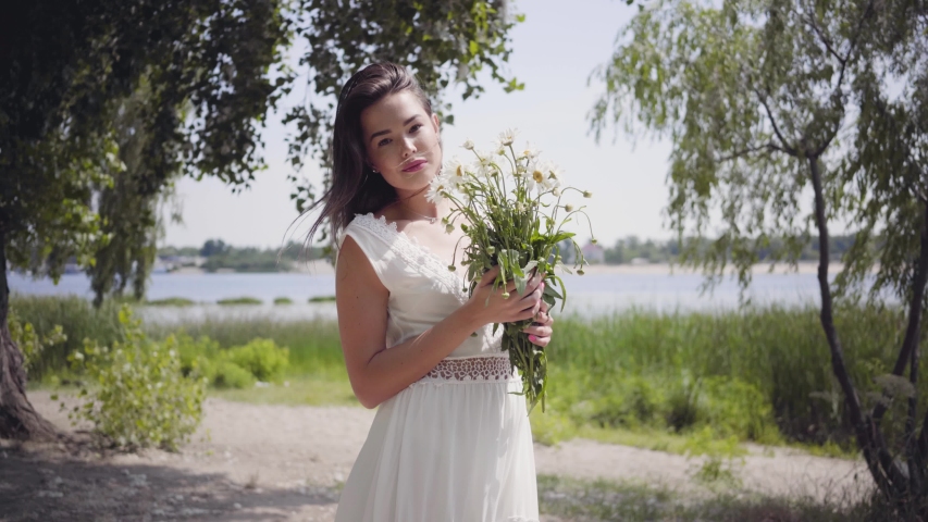 Portrait of cute young girl with long brunette hair wearing a long white summer fashion dress standing in the park. Leisure pretty woman holding a bouquet of chamomile looking at the camera outdoors.