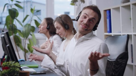 Frustrated young call center operators acting crazy, mad laughing and hilariously typing the keyboard, male operator looks straight to camera, shrugs his shoulders and smiles. Working too hard, no