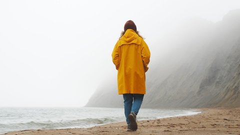 Beautiful european female traveler walks hiking route, enjoy scenic landscape, exploring sea shore, windy foggy weather. Woman wearing yellow raincoat during misty weather. Backpack travel concept Stock Video