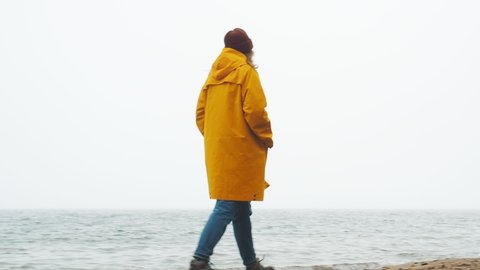 Beautiful european female traveler walks hiking route, enjoy scenic landscape, exploring sea shore, windy foggy weather. Woman wearing yellow raincoat during misty weather. Backpack travel concept วิดีโอสต็อก