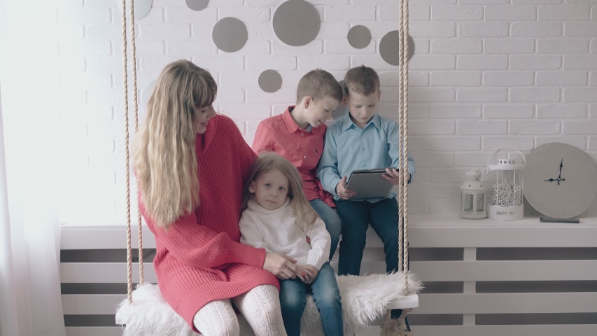 young blond woman and little girl ride on ropes swing two excited boys play video games on modern tablet in room closeup