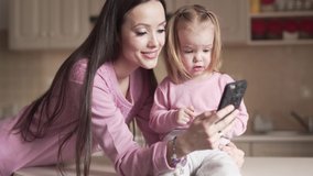 Young beautiful mother is sitting in a kitchen and clicking on a phone with her little cute daughter. They both are smiling and wearing a rose pajamas.