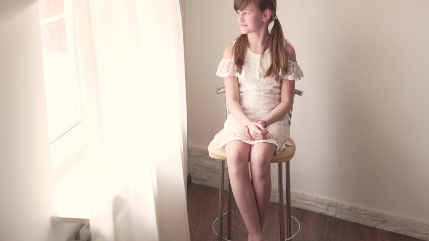 Child Portrait of Girl Sitting in Chair By Window on Daylight in White Dress Moving Her Pigtals to Look Perfetcly and Looks Through Window. Camera Close Up.