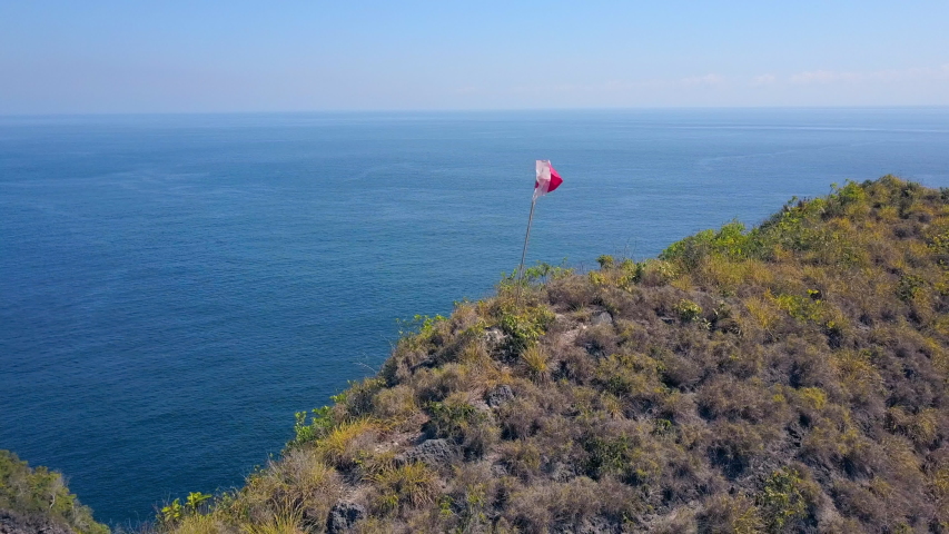 Aerial Backward: Indonesian flag Flying on Ridge Over Bright Blue Ocean - Nusa Penida, Bali