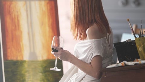 Beautiful woman sits in her bright art studio holding a glass of wine while stroking the canvas with her paintbrush.