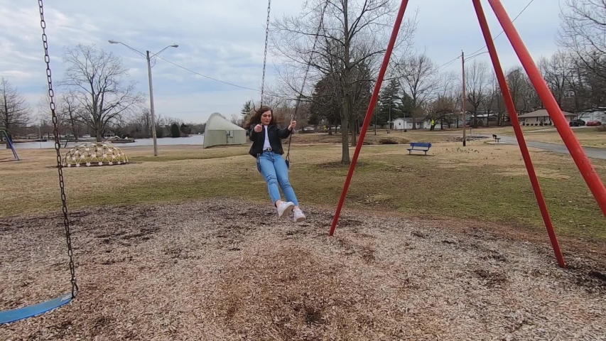 Slow motion footage of a beautiful brunette college teenager swinging in a city park wearing jeans, a white shirt and a black jacket.