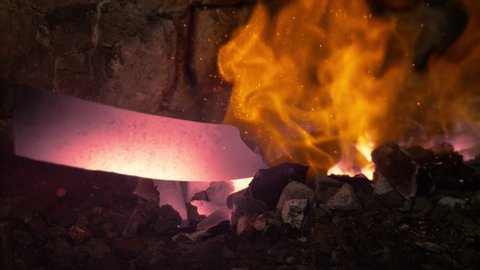 SLOW MOTION, MACRO, DOF: Unrecognizable person forging a blade pulls a hot piece of metal out of the burning ember. Blacksmith pulls a glowing knife blade out of the fire burning inside brick furnace.