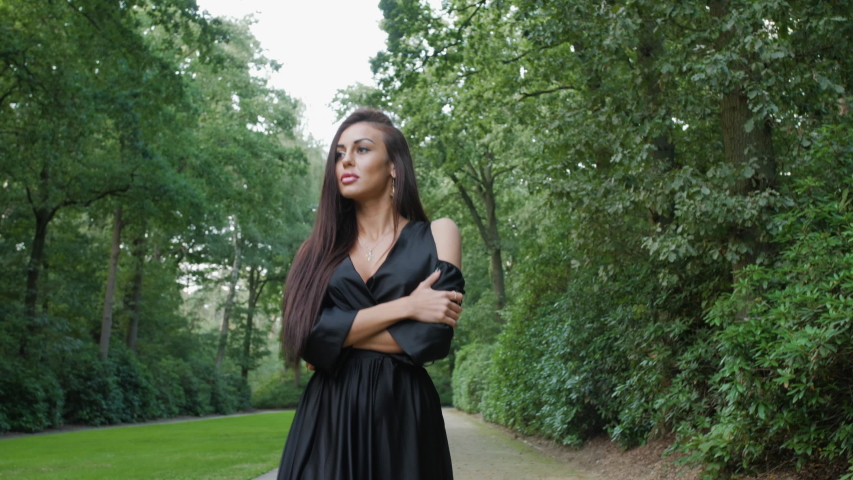 Brunette wearing a black dress and high heels walking in a national park in Belgium, surrounded by trees. Adjusting her hair while looking into the camera and to the side.