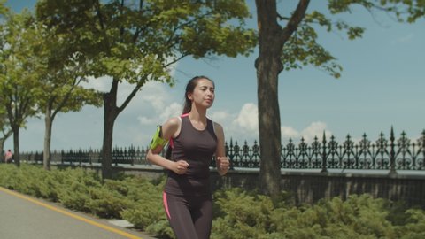 Asian female athlete in sportswear jogging along running path in well-kept city park. Brunette woman jogger with armband running during sport workout outdoors, active, healthy lifestyle concept.