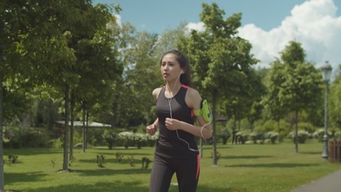 Active female runner with headphones connected to cellphone in armband jogging over green nature of city park in background. Sporty asian woman listening to music during cardio training outdoors.