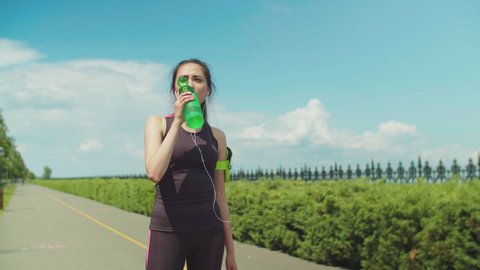 Portrait of chinese female athlete drinking energy drink while running across city park on sunny morning. Sporty woman taking break to drink during training over green nature landscape background.