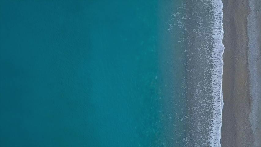 Top view beautiful turquoise waves break on empty pebble beach. Pure calm sea from bird's eye view, drone camera flies over blue waves reaching shore, idyllic bachground for the title