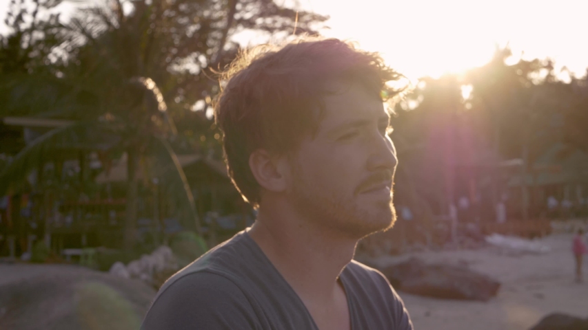 Under the evening sun, a white man with a fresh tan and light beard, sits on a sandy beach and looks out towards the sea.