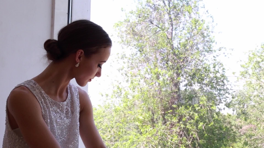 Young tired girl ballerina sitting on a windowsill near a big window in a studio.